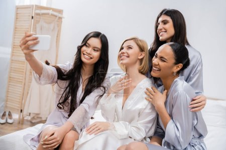 four women, bridal party, joyful blonde bride and her interracial bridesmaids taking selfie together, happiness, silk robes, engagement ring, brunette and blonde, best friends, diversity