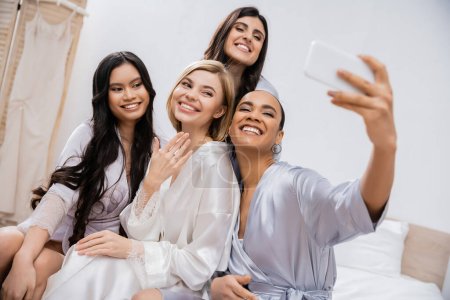 vier Frauen, Brautparty, fröhliche blonde Braut und ihre gemischtrassigen Brautjungfern beim gemeinsamen Selfie, Glück, Seidenroben, Verlobungsring, brünett und blond, beste Freunde, Vielfalt