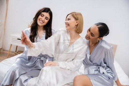 happy blonde bride showing engagement ring to her multicultural bridesmaids, sitting on bed together, bridal shower, silk robes, brunette and blonde women, diversity, multicultural best friends 