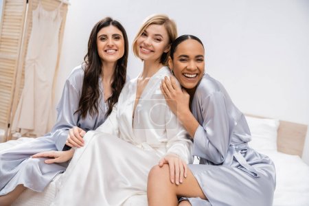 bridal shower, happy blonde bride and her multicultural bridesmaids sitting on bed, showing engagement ring, happiness, silk robes, brunette and blonde, diversity, best friends, three women