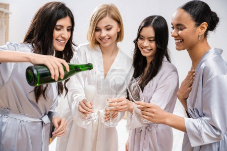 happy woman pouring champagne into glasses near interracial girlfriends, celebration, joyful bride and bridesmaids, brunette and blonde, diversity, bridal shower, best friends, four women 