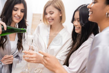 happy woman pouring champagne into glasses near multicultural girlfriends, celebration, cheerful bride and bridesmaids, brunette and blonde, diversity, bridal shower, best friends, four women 