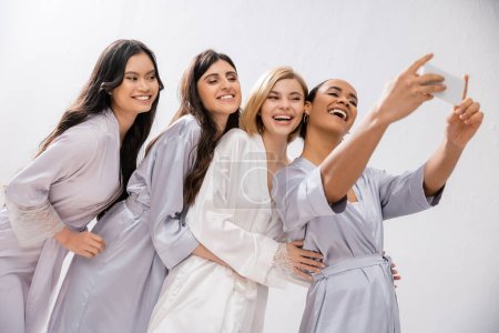 Brautdusche, vier Frauen beim Selfie, glückliche Braut und Brautjungfern in Seidenroben, kulturelle Vielfalt, Spaß zusammen, Freundschaftsziele, brünette und blonde Frauen, Smartphone, digitales Zeitalter