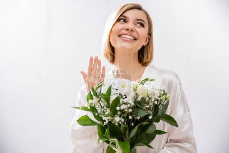 white flowers, happiness, cheerful bride with blonde hair standing in white silk robe and holding bridal bouquet, showing engagement ring, young woman, beautiful, excitement, feminine, blissful 