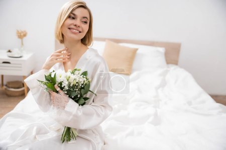 Vorfreude und Glück, fröhliche Braut mit blonden Haaren auf dem Bett sitzend und Brautstrauß haltend, junge Frau im weißen Gewand, schön, Aufregung, feminin, selig, Hochzeitsvorbereitung
