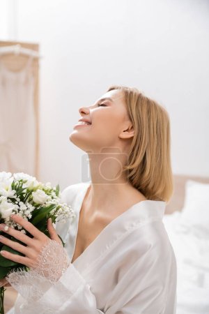 Glück, fröhliche Braut mit blonden Haaren sitzt auf dem Bett und hält Brautstrauß, junge Frau im weißen Gewand, schön, Aufregung, feminin, selig, Hochzeitsvorbereitung, weiße Blumen 