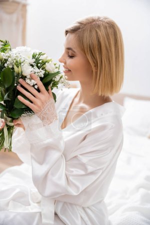 Glück, fröhliche Braut mit blonden Haaren, die auf dem Bett sitzt und weiße Blumen riecht, Brautstrauß, junge Frau im weißen Gewand, schön, Aufregung, feminin, selig, Hochzeitsvorbereitung 