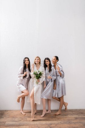 Brautdusche, multikulturelle Frauen mit Gläsern mit Champagner, Braut mit Strauß, die ihren Verlobungsring zeigt, Brautjungfern, Vielfalt, Positivität, weiße Blumen, grauer Hintergrund 