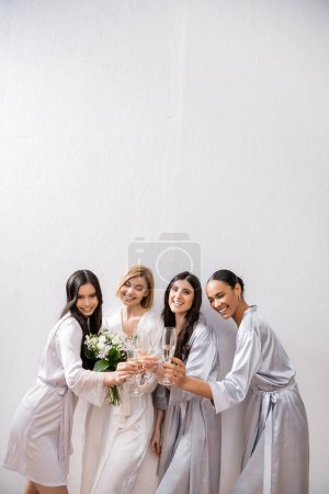 heureux multiculturels copines cliquetis verres avec champagne, mariée avec des fleurs blanches, femmes brunes et blondes, demoiselles d'honneur, diversité, positivité, bouquet nuptial, fond gris 