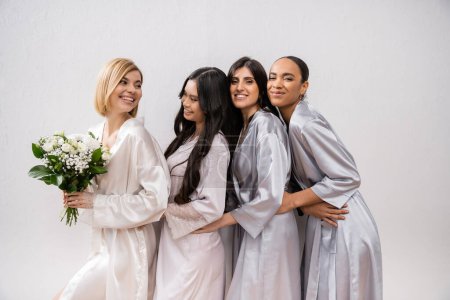 excitement, joyful bride holding bouquet of flowers and standing near cheerful interracial bridesmaids on grey background, racial diversity, silk robes, fashion, brunette and blonde women 