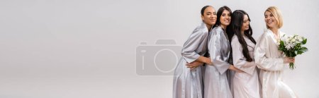 positivity, happy bride holding bouquet of flowers and standing near cheerful interracial bridesmaids on grey background, racial diversity, silk robes, fashion, brunette and blonde, banner 