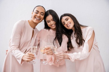 trois demoiselles d'honneur multiculturelles, jolies femmes en robes rose pastel cliquetis verres de champagne sur fond gris, diversité culturelle, mode, célébration, acclamations 
