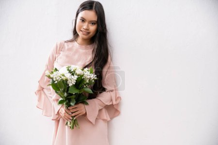 fröhliche Brautjungfer in pastellrosa Kleid mit Blumenstrauß, asiatische Frau mit brünetten Haaren auf grauem Hintergrund, weiße Blumen, besonderen Anlass, Hochzeit, Mode, Lächeln und Freude, Blick in die Kamera 