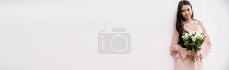 asiatische Brautjungfer in pastellrosa Kleid hält Strauß, glückliche Frau mit brünetten Haaren auf grauem Hintergrund, weiße Blumen, besonderen Anlass, Hochzeit, Mode, Lächeln und Freude, Banner 
