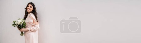 Foto de Ocasión especial, dama de honor alegre en vestido rosa pastel celebración ramo de novia, mujer morena sobre fondo gris, flores blancas, boda, moda, sonrisa y alegría, mirando a la cámara, pancarta - Imagen libre de derechos