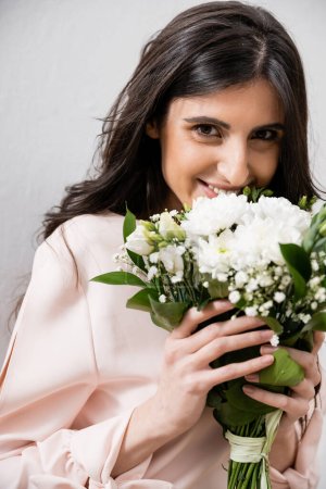 occasion spéciale, heureuse demoiselle d'honneur en robe rose pastel tenant bouquet, femme brune sur fond gris, fleurs blanches, mariage, mode, sourire et joie, en regardant la caméra 