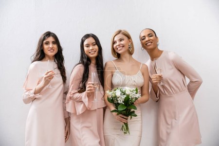 Positivität, glückliche Braut im Brautkleid, Brautstrauß haltend und neben interrassischen Brautjungfern auf grauem Hintergrund stehend, Sektgläser, rassische Vielfalt, Mode, brünette und blonde Frauen 