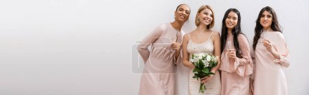 positivity, happy bride in wedding dress holding bridal bouquet and standing near interracial bridesmaids on grey background, champagne glasses, racial diversity, fashion, banner 