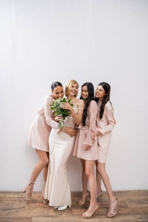 Aufregung, fröhliche multikulturelle Brautjungfern umarmen Braut im Brautkleid, Brautstrauß, grauer Hintergrund, Rassenvielfalt, Mode, brünett und blond, Frauengruppe, weiße Blumen 