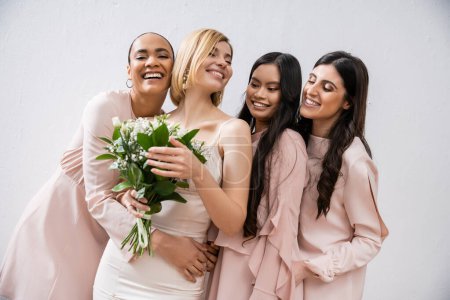 Aufregung, multikulturelle Brautjungfern umarmen glückliche Braut im Brautkleid mit Brautstrauß, grauer Hintergrund, Rassenvielfalt, Mode, brünett und blond, Frauengruppe, weiße Blumen 