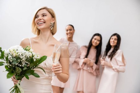cheerful bride in wedding dress holding bridal bouquet and champagne glass, standing near blurred interracial bridesmaids on grey background, happiness, special occasion, blonde and brunette women 
