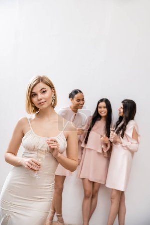 Photo for Beautiful bride in wedding dress holding glass of champagne, standing near blurred interracial bridesmaids on grey background, happiness, special occasion, blonde and brunette women - Royalty Free Image