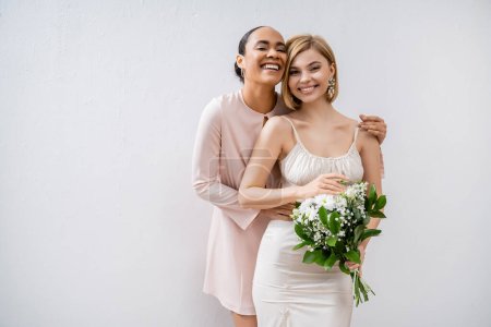 bridal shower, happy bride with bridesmaid, cheerful interracial women, wedding dress and bridesmaid gown, positive african american woman hugging engaged friend on grey background 