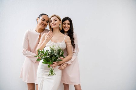 special occasion, bridesmaids hugging bride, friendship goals, grey background, happy multicultural girlfriends, cultural diversity, bridal bouquet, bridal party, blonde and brunette women 