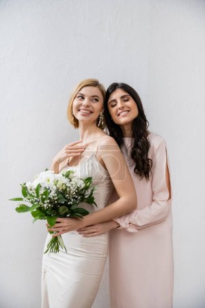 special occasion, bridesmaid hugging bride, friendship goals, grey background, happy girlfriends, bridal bouquet, blonde and brunette women, white flowers, happiness 
