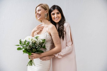 Photo for Special occasion, bridesmaid hugging bride, friendship goals, grey background, cheerful girlfriends, bridal bouquet, blonde and brunette women, white flowers, happiness - Royalty Free Image