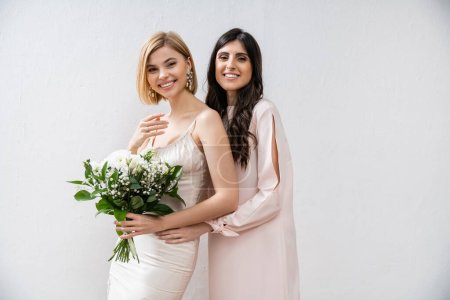 Besonderer Anlass, fröhliche Brautjungfer umarmt Braut, Freundschaftsziele, grauer Hintergrund, glückliche Freundinnen, Brautstrauß, blonde und brünette Frauen, weiße Blumen, Glück 