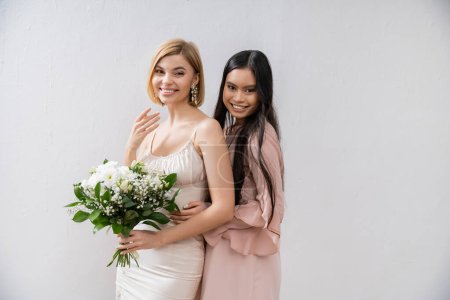 Besonderer Anlass, asiatische Brautjungfer umarmt schöne Braut, Freundschaftsziele, grauer Hintergrund, glückliche Freundinnen, Brautstrauß, blonde und brünette Frauen, weiße Blumen, Positivität 