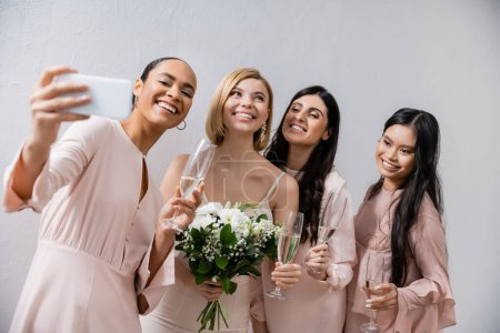 Foto de Cuatro mujeres, novia alegre y sus damas de honor multiculturales tomando selfie juntos, felicidad, copas de champán, ramo de novia, vestido de novia, morena y mujeres rubias - Imagen libre de derechos