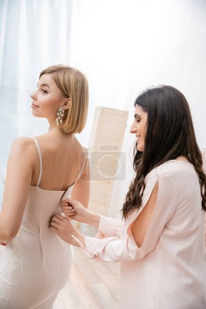 bridesmaid duty, woman zipping white wedding dress of young blonde bride on grey background, fashion, elegance, helping hand, special occasion, happiness, wedding preparations 