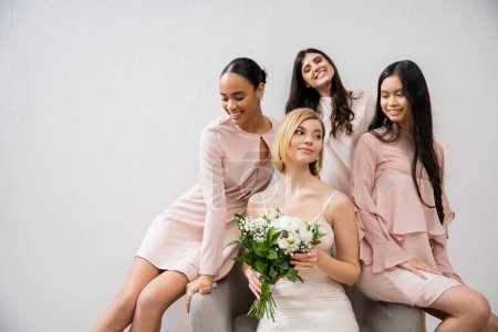 Photo for Wedding photography, four women, joyful bride and bridesmaids, interracial girlfriends, wedding day, cultural diversity, sitting on armchair, grey background, happiness and joy, bridal gown - Royalty Free Image