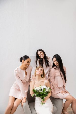 fotografía de boda, diversidad, cuatro mujeres, novia alegre con ramo que muestra su anillo de compromiso cerca de damas de honor, día de la boda, sentado en el sillón, fondo gris, felicidad y alegría 
