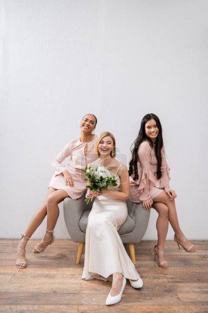 Hochzeitsfotografie, kulturelle Vielfalt, drei Frauen, glückliche Braut mit Strauß und ihre gemischtrassigen Brautjungfern, die auf Sesseln auf grauem Hintergrund sitzen, brünett und blond, Freude, Feier 