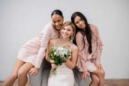 Hochzeitsfotografie, Vielfalt, drei Frauen, glückliche Braut mit Strauß und ihre gemischtrassigen Brautjungfern, die auf Sesseln auf grauem Hintergrund sitzen, brünett und blond, Freude, Feier 