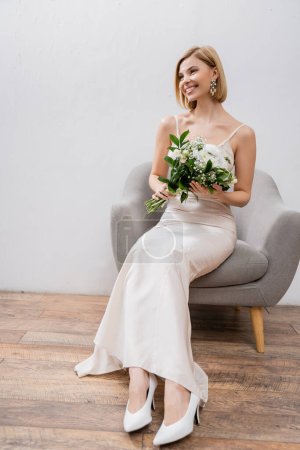 fotografía de boda, ocasión especial, hermosa novia rubia en vestido de novia sentado en sillón y la celebración de ramo sobre fondo gris, flores blancas, accesorios nupciales, felicidad 