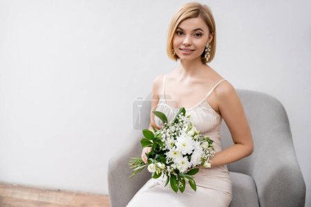ocasión especial, hermosa novia rubia en vestido de novia sentado en sillón y ramo de celebración sobre fondo gris, mirando a la cámara, flores blancas, accesorios nupciales, felicidad, femenino 