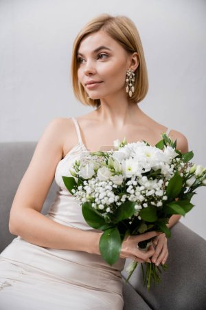 besondere Gelegenheit, schöne und blonde Braut im Hochzeitskleid sitzt im Sessel und hält Strauß auf grauem Hintergrund, weiße Blumen, Brautzubehör, feminin, selig 