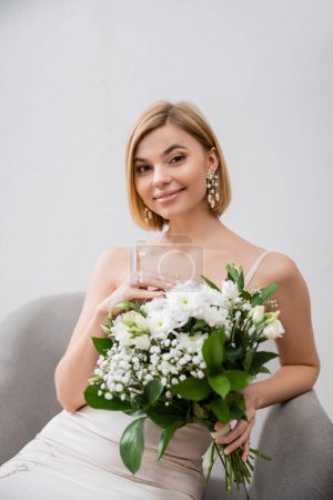 special occasion, beautiful blonde bride in wedding dress sitting in armchair and holding bouquet on grey background, engagement ring, white flowers, bridal accessories, happiness, feminine 