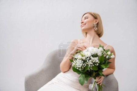 special occasion, happy blonde bride in wedding dress sitting in armchair and holding bouquet on grey background, engagement ring, white flowers, bridal accessories, happiness, feminine 