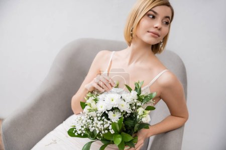 Besonderer Anlass, schöne junge Braut im Brautkleid sitzt im Sessel und hält Blumenstrauß auf grauem Hintergrund, Verlobungsring, weiße Blumen, Brautzubehör, Glück, feminin 