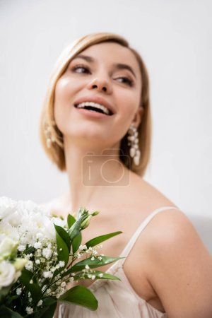 special occasion, beautiful, blonde bride in wedding dress holding bouquet on grey background, white flowers, bridal accessories, happiness, feminine, blissful, looking away, joy 