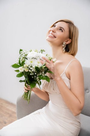 attractive and blonde bride in wedding dress sitting in armchair and holding bouquet on grey background, white flowers, bridal accessories, happiness, special occasion, smiling, feminine, blissful 
