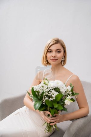 Foto de Novia elegante y rubia en vestido de novia sentado en sillón y ramo de celebración sobre fondo gris, flores blancas, accesorios nupciales, felicidad, ocasión especial, hermoso, femenino, dichoso - Imagen libre de derechos
