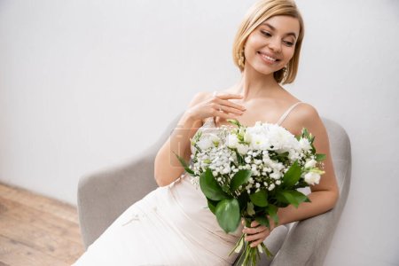 cheerful and elegant bride in wedding dress sitting in armchair and holding bouquet on grey background, white flowers, bridal accessories, happiness, special occasion, beautiful, feminine, blissful 