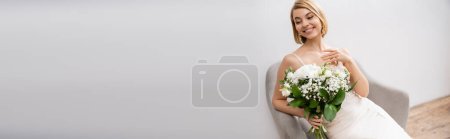 cheerful and blonde bride in wedding dress sitting in armchair and holding bouquet on grey background, white flowers, bridal accessories, happiness, special occasion, beautiful, feminine, banner