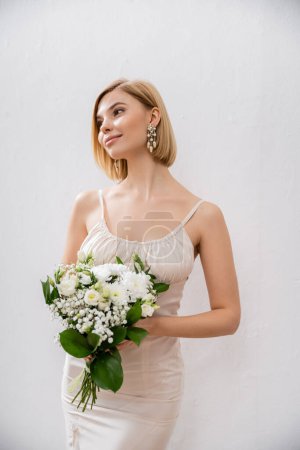 attraktive und blonde Braut im Brautkleid mit Blumenstrauß auf grauem Hintergrund, weiße Blumen, Brautzubehör, Glück, besonderer Anlass, schön, feminin, selig 